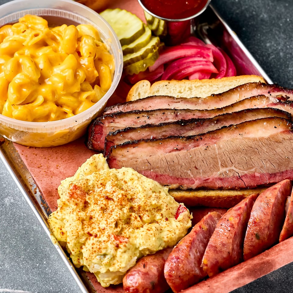 Barbecue platter with sliced brisket, sausage, macaroni and cheese, potato salad, pickles, onions, toast, and barbecue sauce.