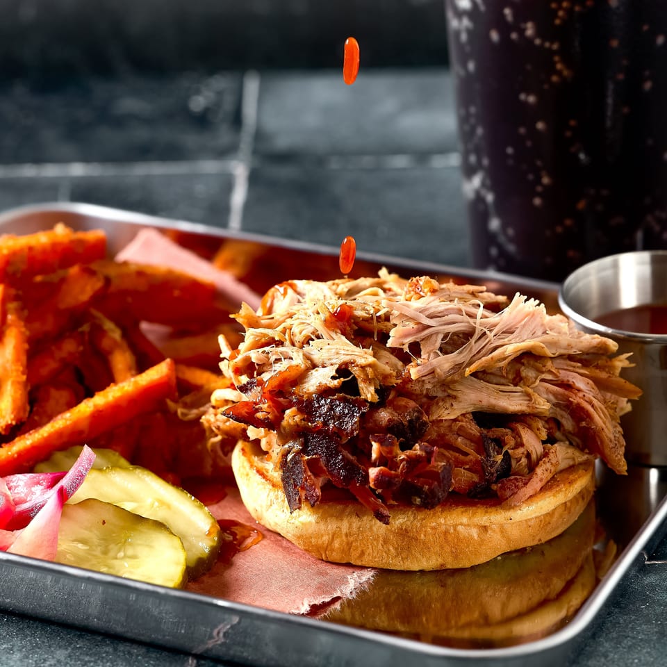 Loaded pulled-meat sandwich with crispy edges, fries, pickles, and sauce in a serving tray beside a cola.