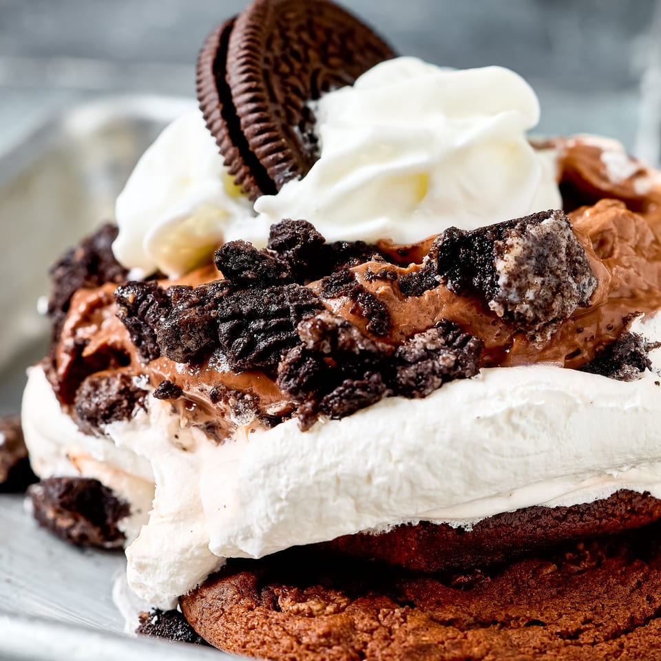 Close-up of a chocolate cake slice layered with whipped cream, chocolate frosting, cookie crumbs, and a chocolate sandwich cookie.