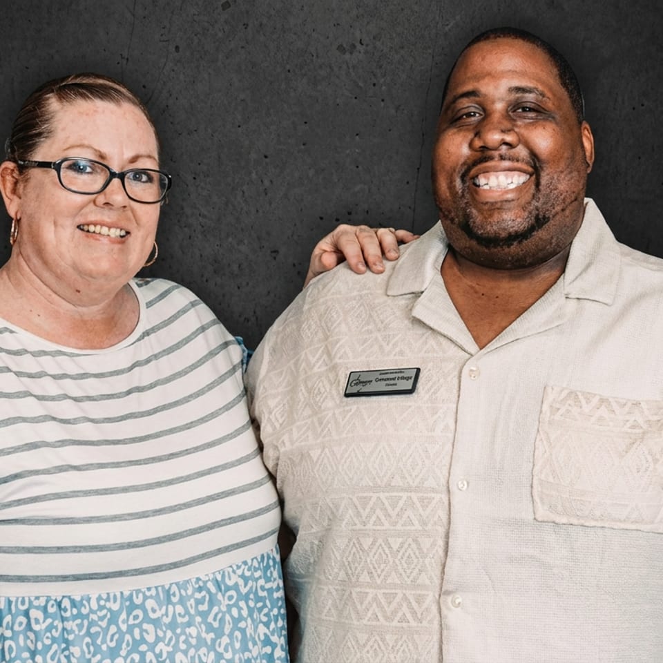 Smiling woman and man pose together against a dark backdrop, with the man wearing a name badge and patterned shirt.