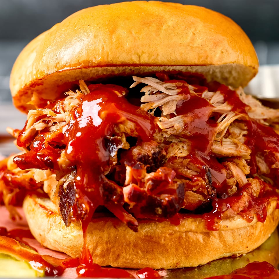 Close-up of a saucy pulled chicken sandwich with shredded meat and vegetables on a toasted bun.